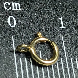 Pode incluir: Um fecho de metal dourado com um anel redondo, medindo aproximadamente 1 cm de di&acirc;metro. O fecho &eacute; mostrado contra um fundo preto com uma r&eacute;gua marcada em cent&iacute;metros.