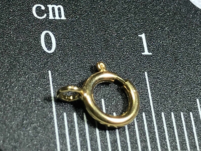 Pode incluir: Um fecho de metal dourado com um anel redondo, medindo aproximadamente 1 cm de di&acirc;metro. O fecho &eacute; mostrado contra um fundo preto com uma r&eacute;gua marcada em cent&iacute;metros.