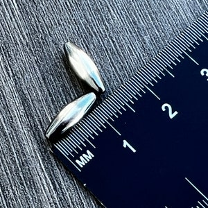 May include: Two silver-coloured metal beads, measuring approximately 1 cm in length, are placed next to a black ruler with white markings. The ruler shows measurements in millimetres.