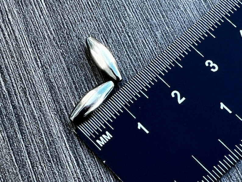 May include: Two silver-coloured metal beads, measuring approximately 1 cm in length, are placed next to a black ruler with white markings. The ruler shows measurements in millimetres.
