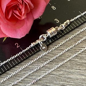 May include: A silver chain necklace featuring a cylindrical magnetic clasp and a small heart-shaped tag stamped with 925. The necklace is positioned against a black ruler showing measurements in inches, with the clasp spanning approximately 0.5 inches.