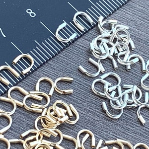 Op de afbeelding: Een close-up van twee stapels kleine, U-vormige sprongringen van metaal. De ene stapel is goud en de andere is zilver. De ringen zijn in een willekeurig patroon gerangschikt. Een liniaal is zichtbaar op de achtergrond.