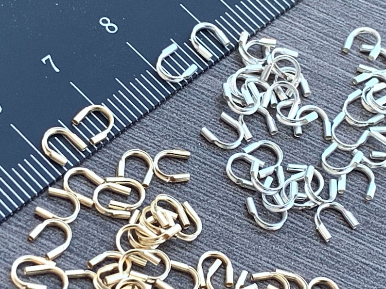 Op de afbeelding: Een close-up van twee stapels kleine, U-vormige sprongringen van metaal. De ene stapel is goud en de andere is zilver. De ringen zijn in een willekeurig patroon gerangschikt. Een liniaal is zichtbaar op de achtergrond.