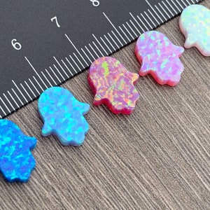 May include: Five hamsa hand charms made of opal in various colors, including blue, pink, and white. The charms are arranged in a row on a black and brown surface.