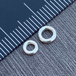 Puede incluir: Dos anillos de salto de plata, uno más grande que el otro, se muestran junto a una regla. El anillo de salto más grande tiene un diámetro de aproximadamente 1,2 cm. El anillo de salto más pequeño tiene un diámetro de aproximadamente 0,6 cm.