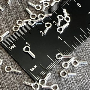 May include: A close-up of silver-toned metal eye pins, measuring approximately 2.5 cm in length, scattered on a wooden surface. A ruler is visible in the background, with markings in inches and millimetres.