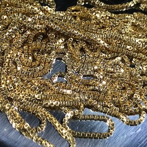 Puede incluir: Primer plano de un collar de cadena de oro con un patrón de cadena de caja. La cadena está hecha de pequeños eslabones cuadrados que se entrelazan.