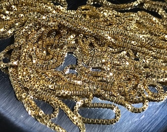 May include: A close-up of a gold chain necklace with a box chain pattern. The chain is made of small, square links that are interlocked together.