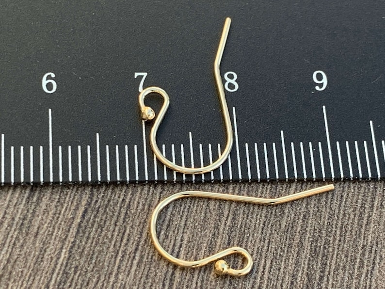 May include: Two gold-toned earring hooks with a small ball at the end of each hook. The hooks are measured against a ruler with markings from 6 to 9.