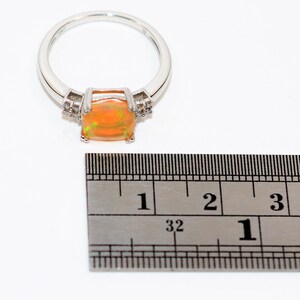 May include: A silver ring with a large orange opal gemstone and smaller clear gemstones on the band. The ring is measured against a ruler, showing the stone is about 1 inch wide.