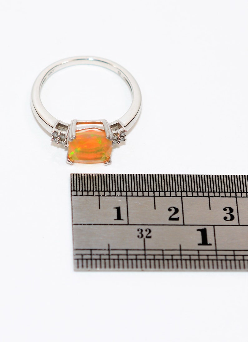 May include: A silver ring with a large orange opal gemstone and smaller clear gemstones on the band. The ring is measured against a ruler, showing the stone is about 1 inch wide.