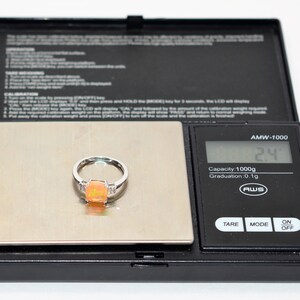 May include: A silver ring with a large orange opal gemstone and smaller clear gemstones on the band. The ring is on a digital scale that reads 2.4 grams.