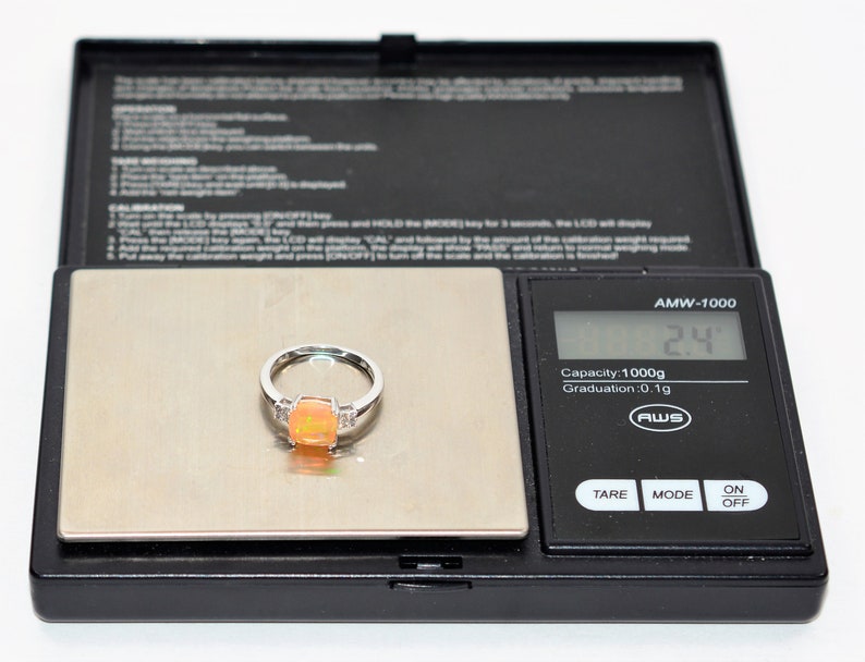 May include: A silver ring with a large orange opal gemstone and smaller clear gemstones on the band. The ring is on a digital scale that reads 2.4 grams.