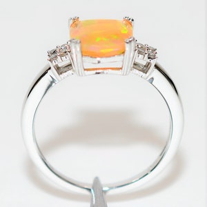 May include: A silver ring with a large, square-shaped opal gemstone in the center. The opal is a pale orange color with flashes of green and yellow. The ring is set with smaller diamonds on either side of the opal.