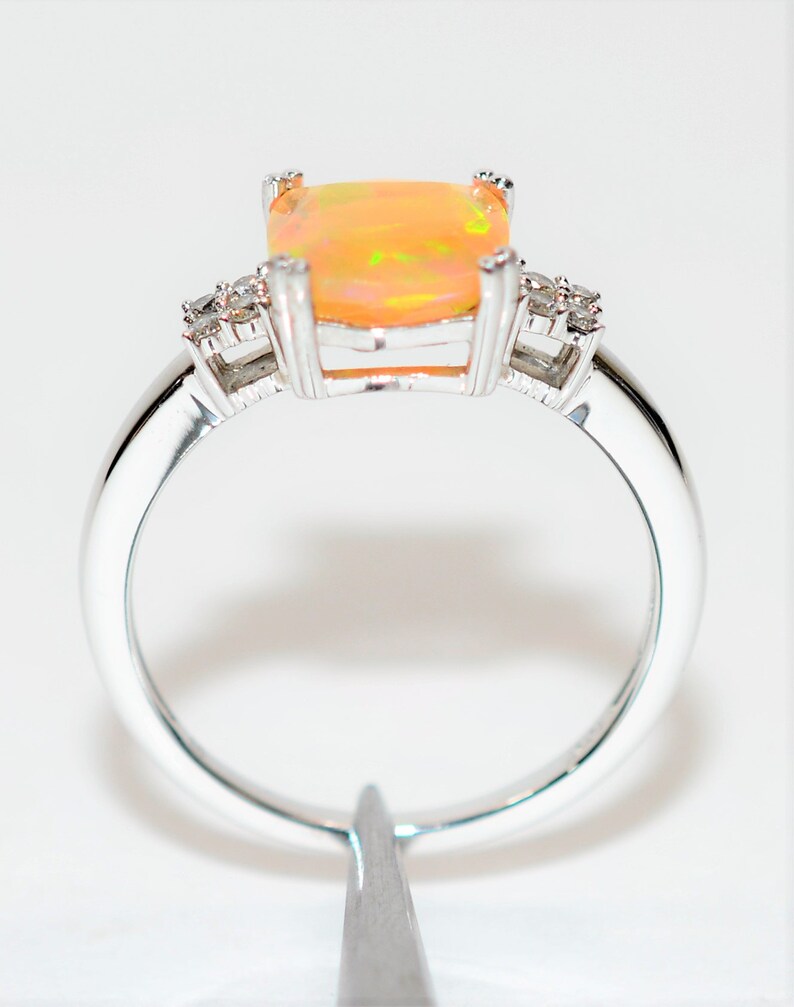 May include: A silver ring with a large, square-shaped opal gemstone in the center. The opal is a pale orange color with flashes of green and yellow. The ring is set with smaller diamonds on either side of the opal.