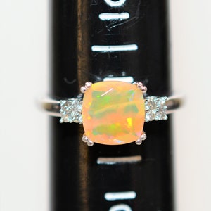 May include: A silver ring with a large, light pink opal gemstone surrounded by smaller clear gemstones. The ring is on a black ruler with white markings.
