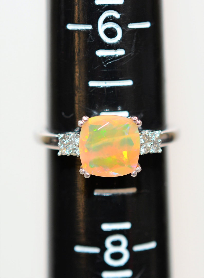 May include: A silver ring with a large, light pink opal gemstone surrounded by smaller clear gemstones. The ring is on a black ruler with white markings.