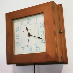 May include: A wooden framed clock with a white face and blue numbers. The clock hands are black and the clock is plugged in.
