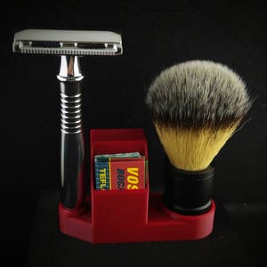 May include: A red plastic stand with a chrome safety razor, a shaving brush with black bristles, and a stack of razor blades. The stand has the words "VOS" and "BOCK" printed on it.