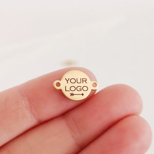 Könnte beinhalten: Goldfarbener Metallanhänger mit runder Form und zwei Sprungringen. Der Anhänger trägt den Text "YOUR LOGO" eingraviert mit einem Pfeil, der nach rechts zeigt.