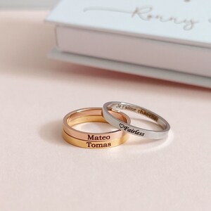 May include: Three gold rings stacked on top of each other. The top ring is silver and engraved with the text "Je t'aime cherie" and a heart symbol followed by the word "Fairless". The middle ring is engraved with the name "Mateo" and the bottom ring is engraved with the name "Tomas".