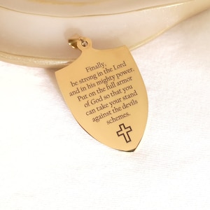 May include: Gold-toned shield pendant necklace with a cross at the bottom. The shield has an inscription that reads: "Finally, be strong in the Lord and in his mighty power. Put on the full armor of God so that you can take your stand against the devils schemes."