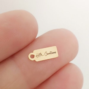 May include: A small, gold-colored tag charm with a handle-like design on the left side. The tag has the text "Hm Creations" in a delicate script. The charm is held in a person's hand against a white background.