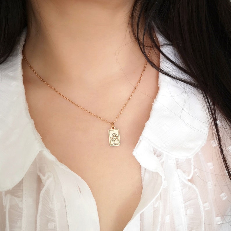May include: A gold chain necklace with a small rectangular pendant featuring a tarot card design. The card depicts a figure sitting on a throne with a crown on their head.
