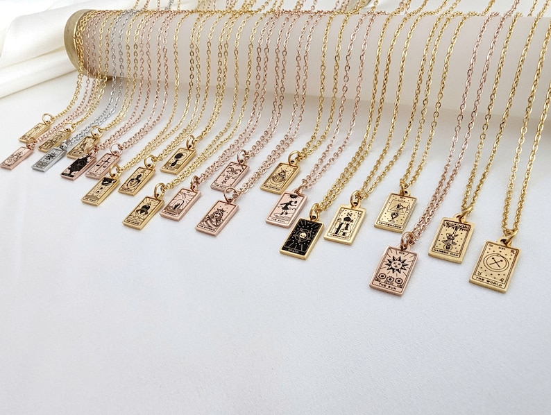 May include: A collection of gold, silver, and rose gold necklaces with small rectangular pendants featuring tarot card symbols. The pendants are engraved with various tarot card symbols, including the sun, the moon, and the world.