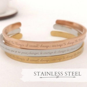 Peut inclure: Trois bracelets en acier inoxydable empilés avec l'inscription "The things I cannot change; courage to change the things" en anglais et en français. Les bracelets sont en or rose, argent et or.