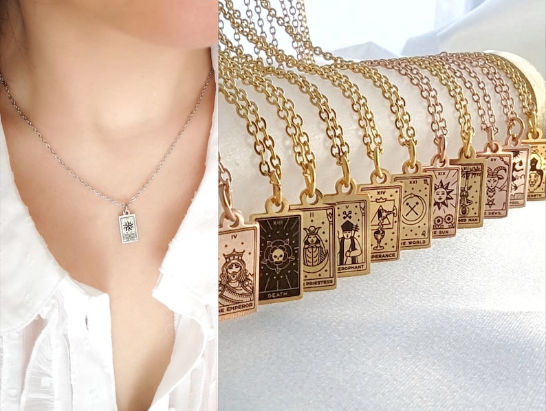 May include: A silver necklace with a small rectangular pendant featuring a star design. The pendant is hanging from a delicate chain. The necklace is layered over a white shirt. In the background, a collection of gold necklaces with rectangular pendants featuring tarot card designs are displayed.