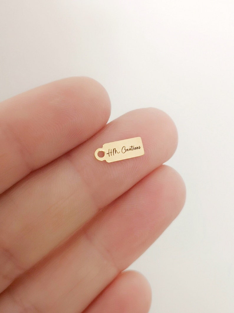 May include: A small, gold-colored tag charm with a handle-like design on the left side. The tag has the text "Hm Creations" in a delicate script. The charm is held in a person's hand against a white background.