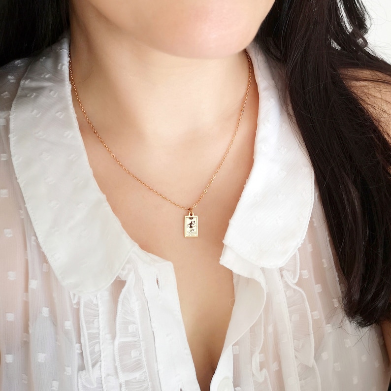 May include: A gold chain necklace with a small rectangular pendant featuring a stylized figure. The necklace is worn by a person with long dark hair and a white blouse.