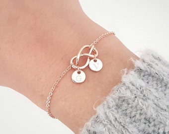 Pode incluir: Um bracelete de nó infinito em ouro rosa com dois pingentes personalizados. Os pingentes são redondos e têm as letras "G" e "V" gravadas.