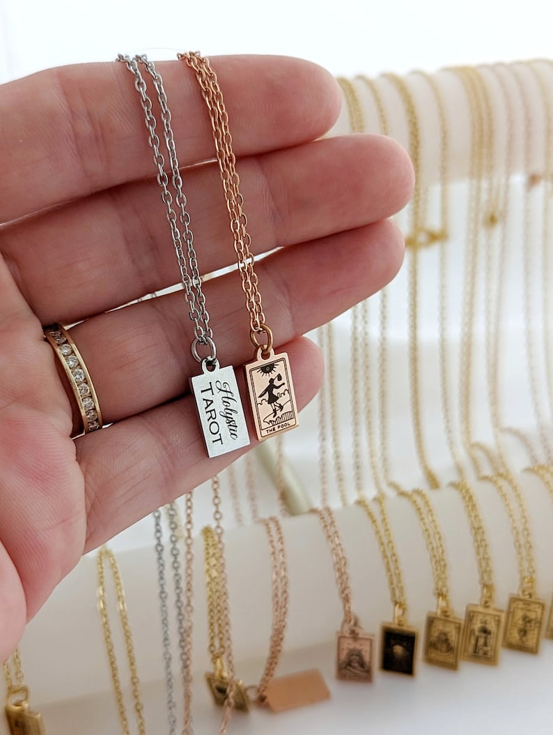 May include: Two silver and gold necklaces with a tarot card pendant. The silver necklace has a rectangular pendant with the text "Holygrail Tarot". The gold necklace has a rectangular pendant with a tarot card image of "The Fool".