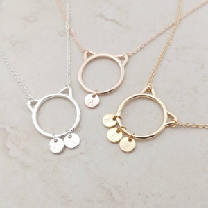 Peut inclure: Trois colliers en or avec des pendentifs en forme de chat et des breloques. Les colliers sont superposés et les breloques sont gravées des lettres A, K, B et F.