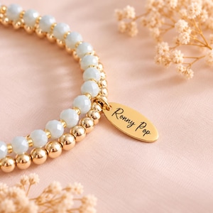 May include: A stretch bracelet featuring alternating sections of faceted pale blue beads and polished gold-tone metallic beads. An oval-shaped gold-tone metal charm is attached to the bracelet, engraved with the black script text Ronny Pop.