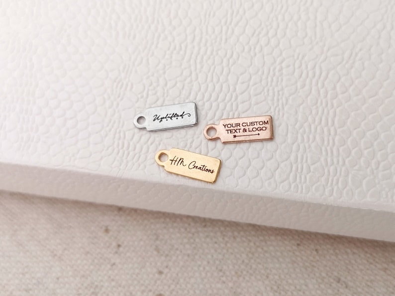 May include: Three small rectangular metal tags with a hole at one end. One tag is silver with the word "Uplifted" in script. Another is rose gold with "YOUR CUSTOM TEXT & LOGO" and an arrow. The third is gold with "HM Creations".