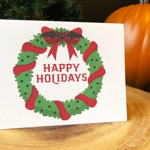 May include: A white card with a red and green wreath with a black bat with red eyes and a spider web. The wreath has a red bow and the text "HAPPY HOLIDAYS" in black.