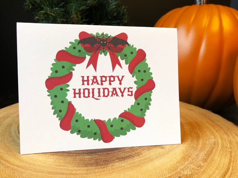 May include: A white card with a red and green wreath with a black bat with red eyes and a spider web. The wreath has a red bow and the text "HAPPY HOLIDAYS" in black.