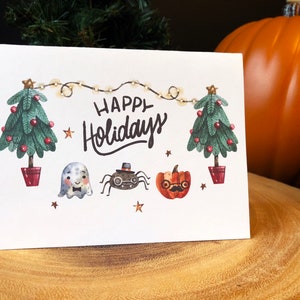 May include: A white card with the text "Happy Holidays" in black cursive lettering. The card is decorated with two green Christmas trees, a ghost, a spider wearing a top hat, and a pumpkin with glasses and a mustache. The trees are decorated with red ornaments and white lights.