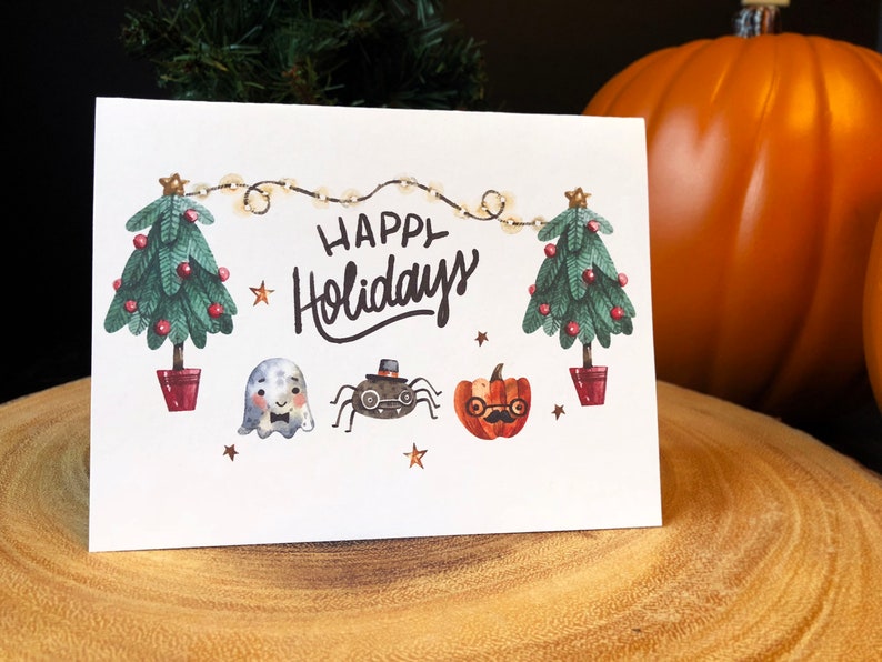 May include: A white card with the text "Happy Holidays" in black cursive lettering. The card is decorated with two green Christmas trees, a ghost, a spider wearing a top hat, and a pumpkin with glasses and a mustache. The trees are decorated with red ornaments and white lights.