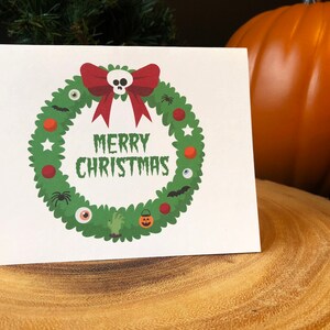 May include: A white card with a green wreath decorated with Halloween-themed ornaments, including a skull, eyeballs, bats, spiders, and a pumpkin. The wreath has a red bow and the text "Merry Christmas" in black.