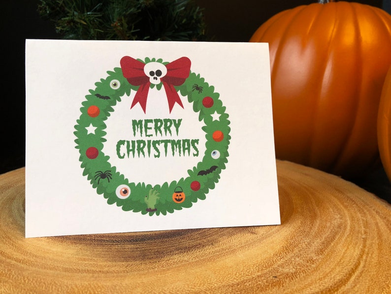 May include: A white card with a green wreath decorated with Halloween-themed ornaments, including a skull, eyeballs, bats, spiders, and a pumpkin. The wreath has a red bow and the text "Merry Christmas" in black.