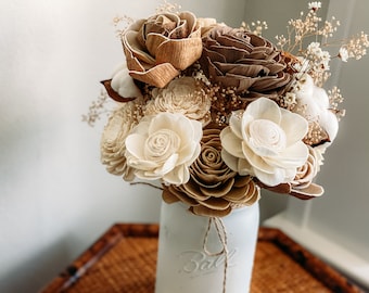 May include: A bouquet of dried flowers in shades of brown and white, arranged in a white painted mason jar. The flowers are made of wood and include roses, peonies, and other varieties. The jar is tied with twine and sits on a woven placemat.