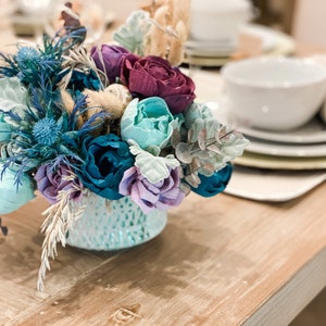 May include: A teal ceramic vase filled with a bouquet of faux flowers. The flowers are in shades of teal, purple, and blue. The vase has a textured surface.