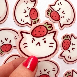 May include: A collection of stickers featuring a cartoon cat with a strawberry on its head. The stickers have a white background with a pink outline and are in various poses. The cat has a grumpy expression.
