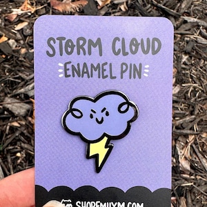 Może przedstawiać: Fioletowa emaliowana przypinka z kreskówkową chmurą burzową z żółtym błyskawicą. Przypinka zawiera napis "STORM CLOUD ENAMEL PIN" i "SHOPEMILYMM.COM".