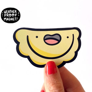 K&ouml;nnte beinhalten: Ein gelber Cartoon-Pierogi mit einem l&auml;chelnden Gesicht, schwarzem Umriss und dem Text "WEATHER PROOF MAGNET!" in einem schwarzen Kreis.