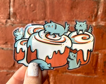 Könnte beinhalten: Ein Aufkleber mit einer Zimtschnecke mit weißem Frosting und einem braunen Zimt-Wirbel. Vier kleine blaue Comic-Katzen sitzen auf der Schnecke, eine Katze sitzt oben auf der Schnecke.